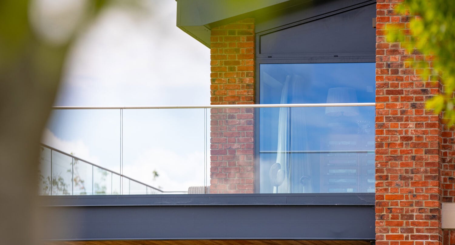 Modern brick house with large blue-tinted windows, bifold doors, and a glass balcony railing, partially obscured by green tree branches in the foreground. The architecture features clean lines and contemporary design elements.