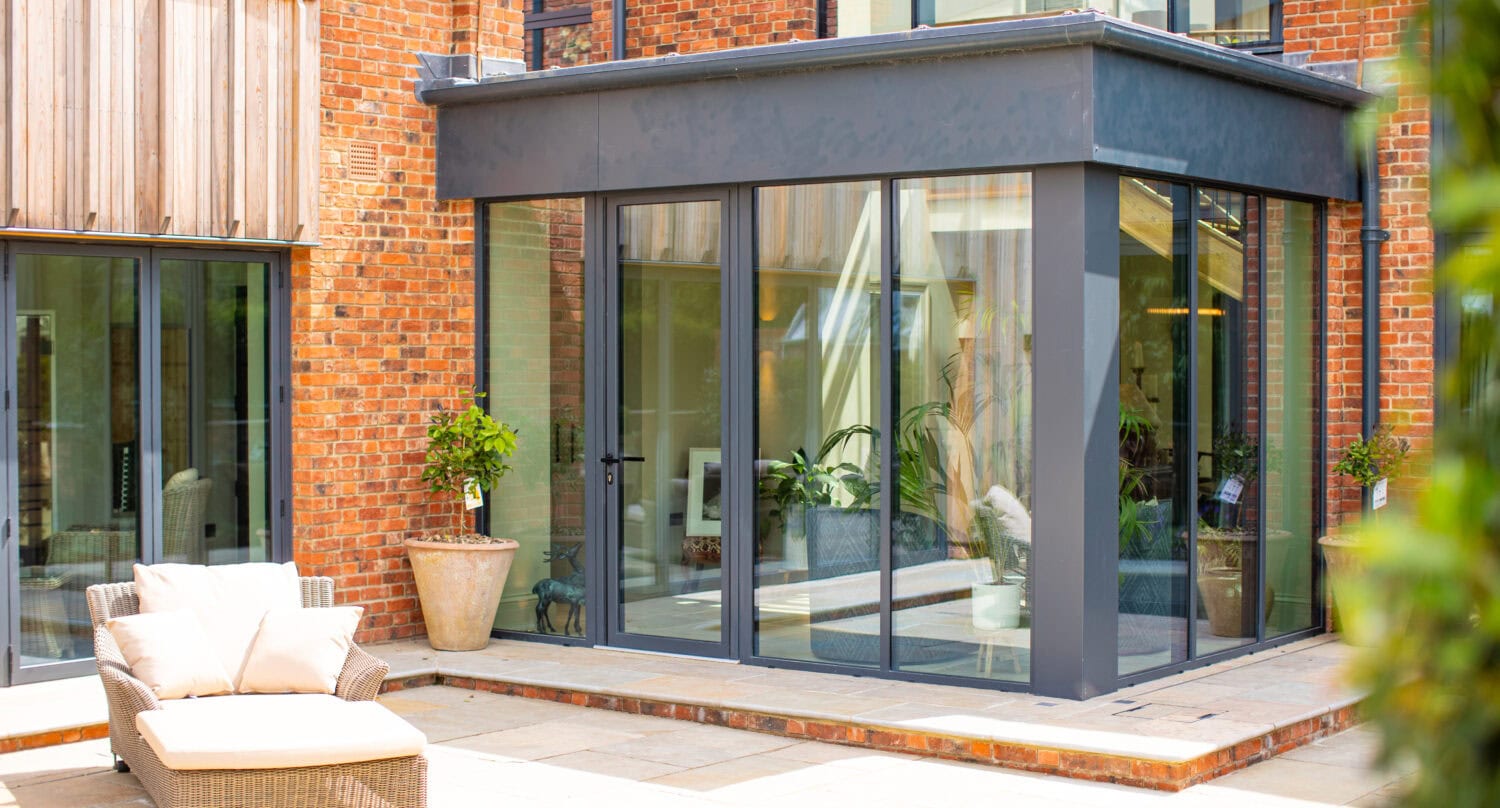 Modern patio area with a cushioned lounge chair in front of a house featuring large glass windows, bifold doors, red brick walls, and potted plants near the entrance.