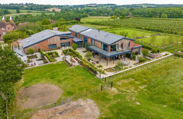 Aerial view of a large, modern house with bifold doors opening to expansive gardens and lawn, surrounded by green fields and trees in a rural area under a cloudy sky.