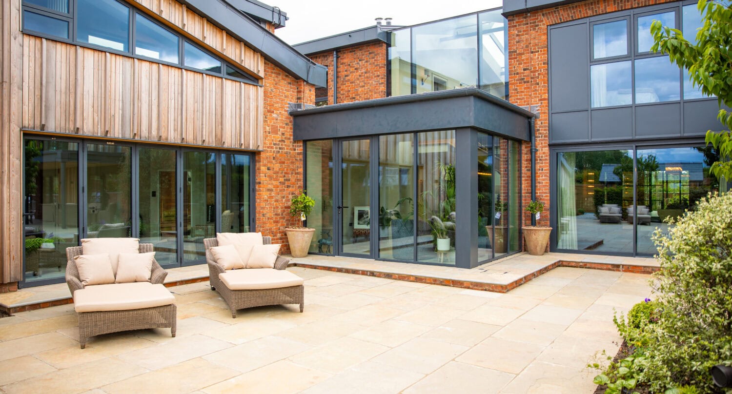 Modern house with large windows, a mix of brick and wood exterior, and spacious stone patio featuring sliding doors, two cushioned lounge chairs, and potted plants. Lush greenery surrounds the outdoor area.