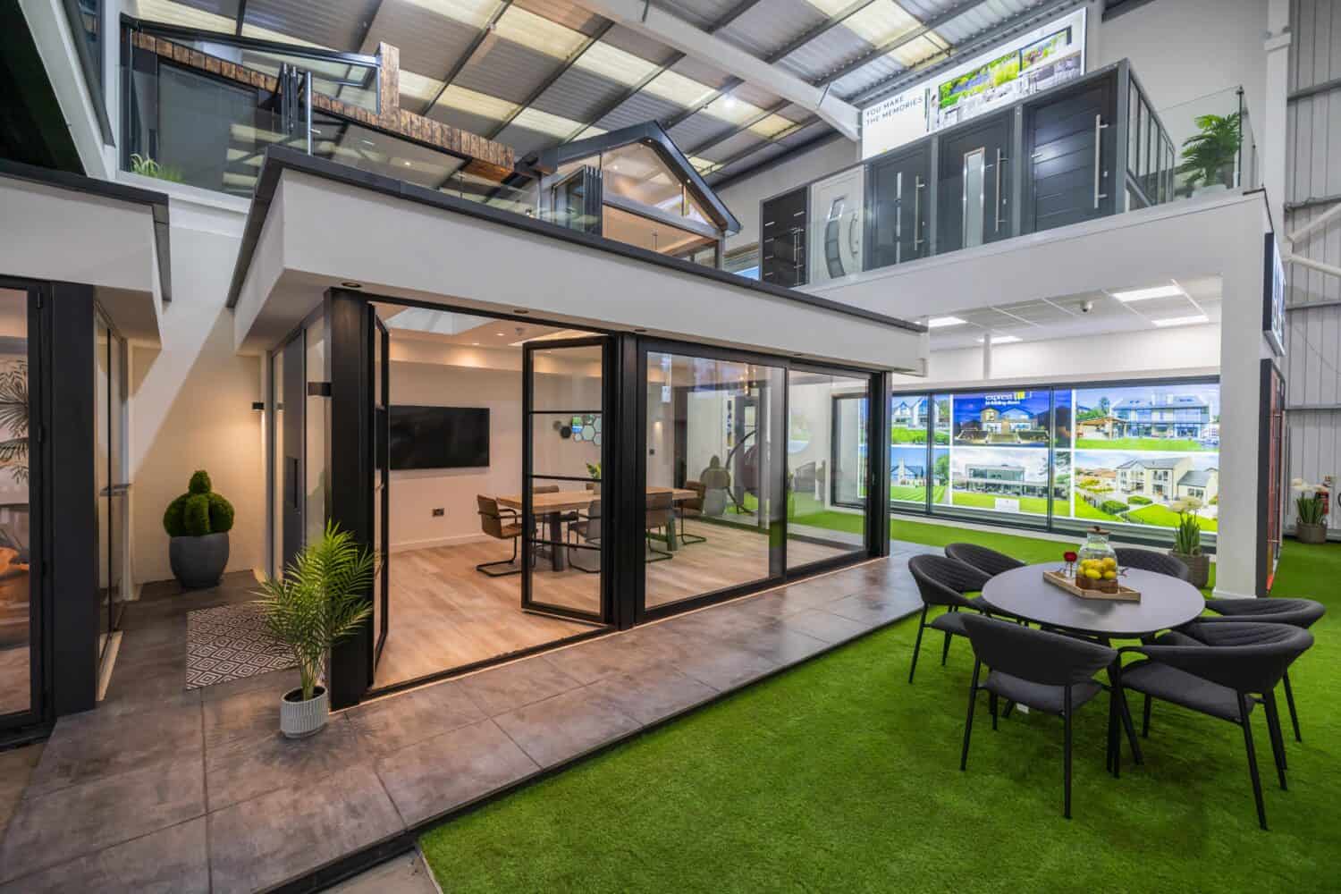 Modern indoor showroom featuring a two-level model home with bespoke glass solutions, including striking glass walls and sliding doors, a dining area inside, and an outdoor seating area on artificial grass. Bright lighting and various home displays are visible in the background.