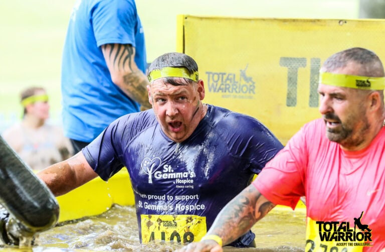 Two men wearing headbands and muddy shirts tackle an outdoor obstacle course, climbing through a water-filled barrier. One man sports a purple St. Gemma’s Hospice shirt; the other wears pink. Bespoke glass solutions banners are visible in the background.