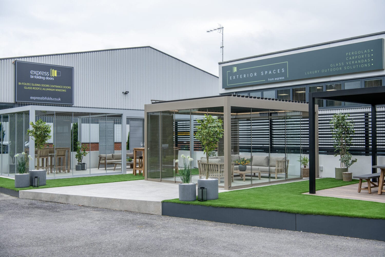 Outdoor showroom featuring modern pergolas, stylish patio furniture, potted plants, artificial grass, and bifold doors outside a commercial building with signs for Express Bi-folding Doors and Exterior Spaces.