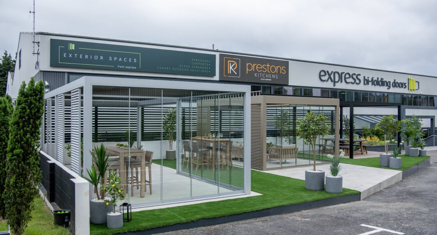 A modern showroom exterior features glass-walled rooms, bifold doors, outdoor seating, potted plants, and signage for Exterior Spaces, Prestons Kitchens, and Express Bi-folding Doors under a cloudy sky.