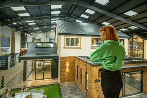 A woman with red hair wearing a bright green shirt stands on a balcony overlooking a modern indoor exhibition space featuring model homes with glass roofing and people seated at a table below.