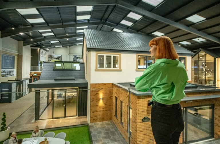A woman with red hair wearing a bright green shirt stands on a balcony overlooking a modern indoor exhibition space featuring model homes with glass roofing and people seated at a table below.