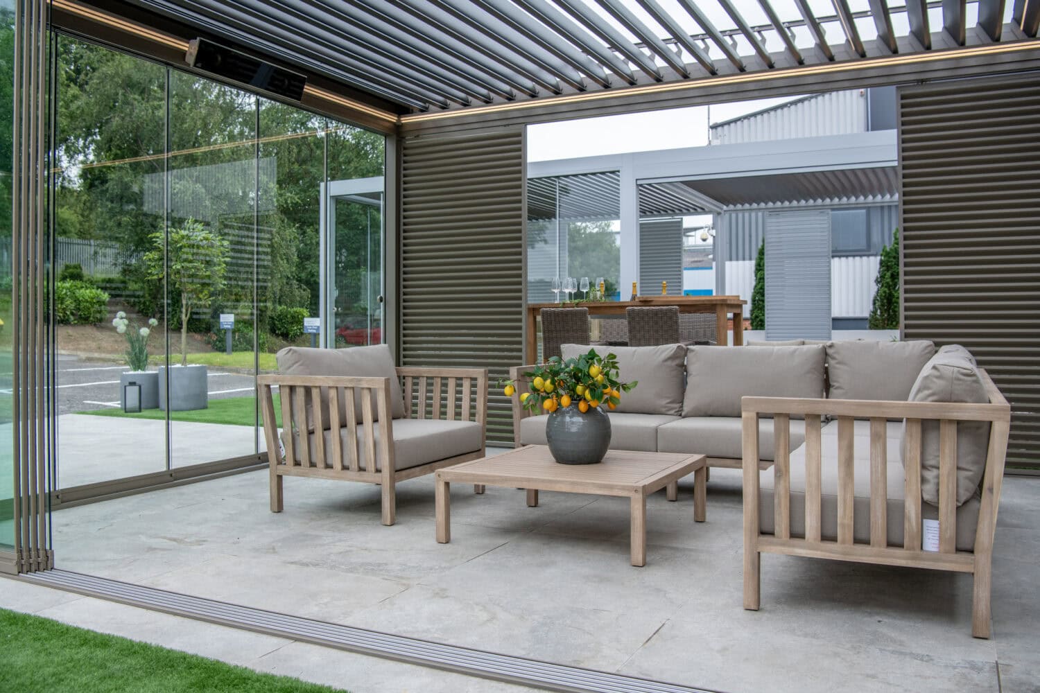 Modern outdoor patio with beige cushioned wooden furniture, a coffee table with a potted plant, and glass walls. Sliding doors open to a dining area and stunning views of the contemporary house and garden.