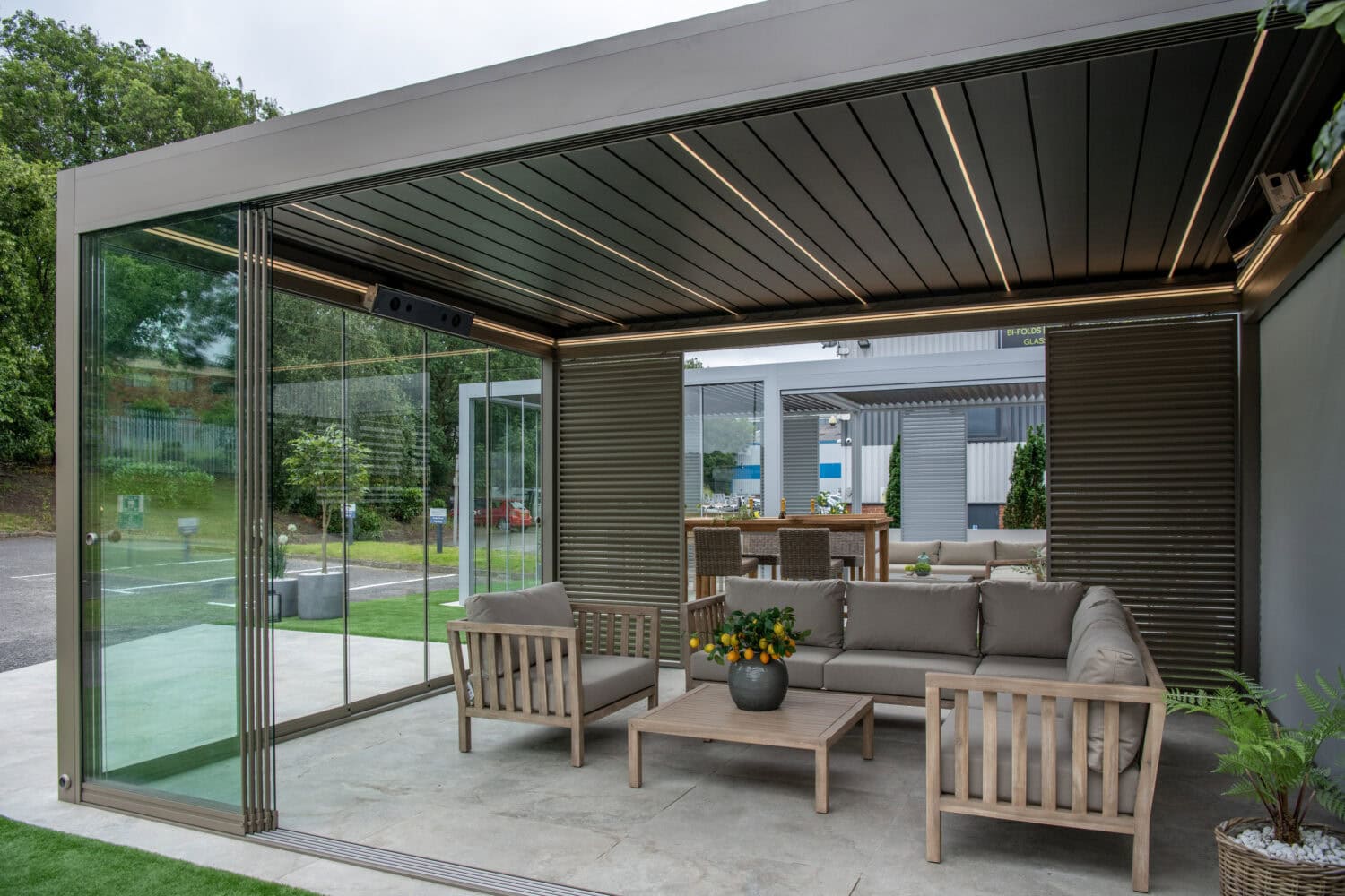 Modern outdoor patio with a covered seating area, featuring bespoke glass solutions like sliding doors, wooden furniture with gray cushions, a coffee table with a plant, and a view of another seating area and greenery outside.