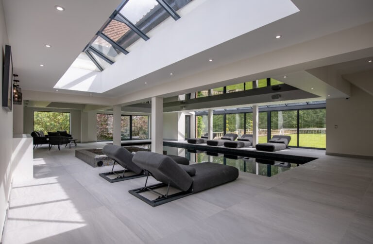 Modern indoor pool area with several black lounge chairs, large windows, skylights, and bespoke glass solutions offering a seamless view of the green garden outside. The bright, spacious space features sleek, contemporary design elements.
