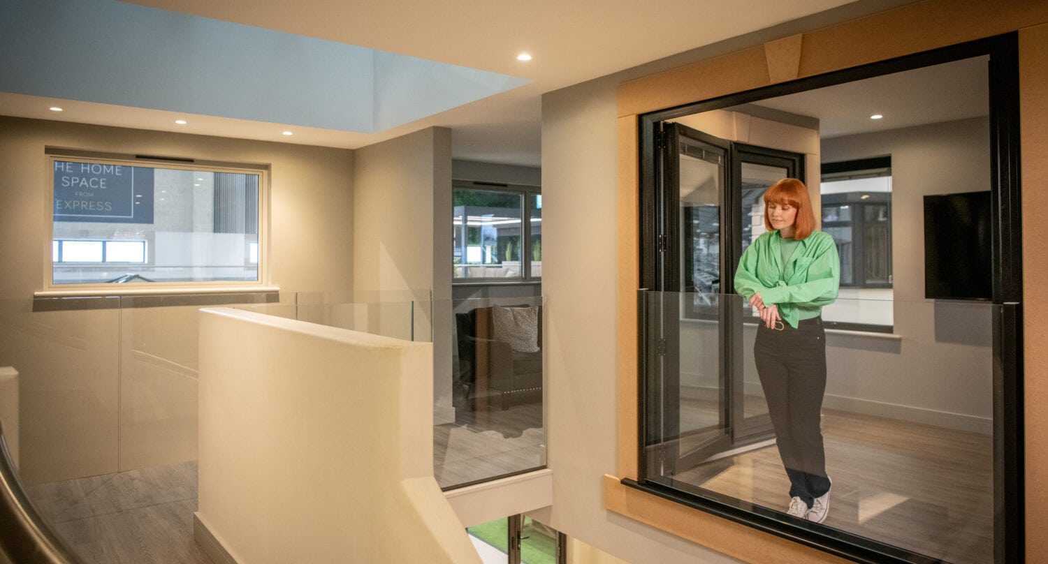 A woman with red hair, wearing a green shirt and jeans, stands inside a glass-walled room featuring bifold doors, overlooking a modern, well-lit interior hallway with clean lines and neutral colors.