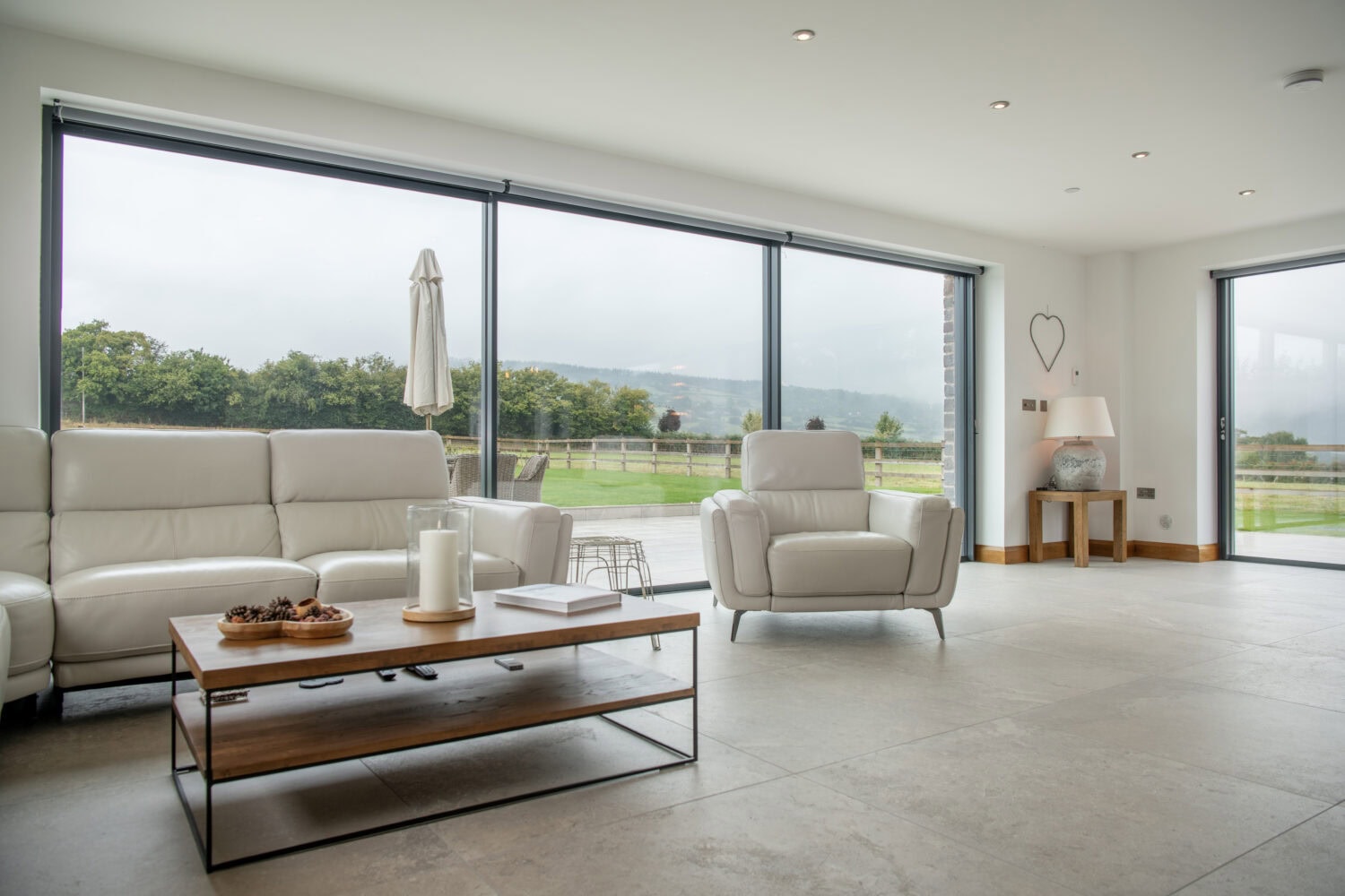Modern living room with light-colored sofas and armchair, large bifold doors and floor-to-ceiling windows overlooking a patio and green landscape, plus a wooden coffee table adorned with candles and a decorative tray.