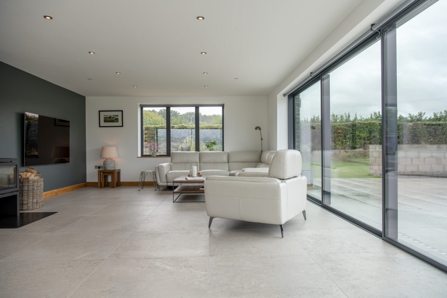 Modern living room with large floor-to-ceiling windows and sliding doors, white leather sofas, a TV, and a lamp. The space is bright with outdoor views and neutral-colored tile flooring.