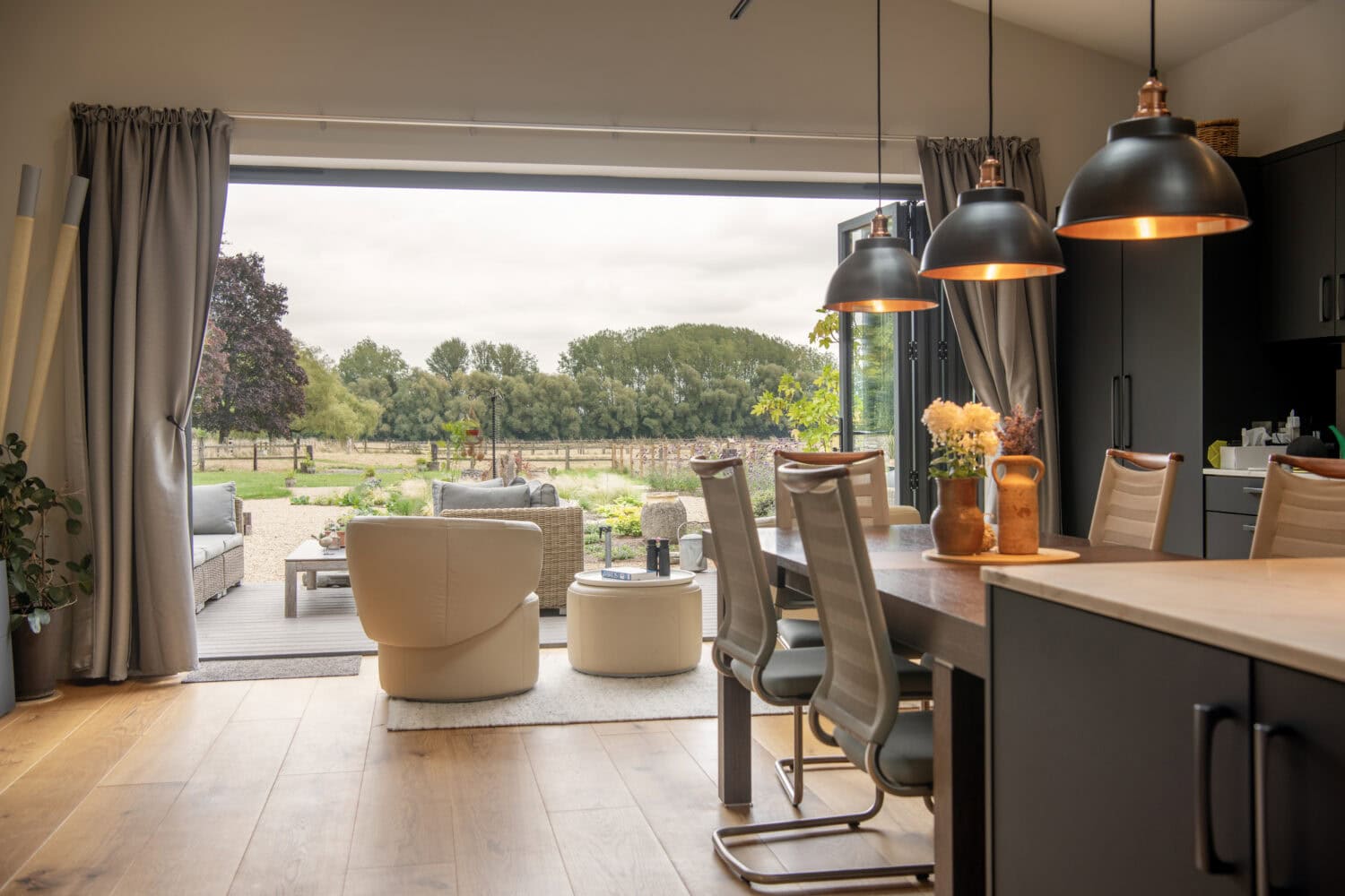 Modern open-plan kitchen and dining area with pendant lights, leading to a cozy patio with outdoor seating; large sliding doors reveal a scenic garden and trees beyond.
