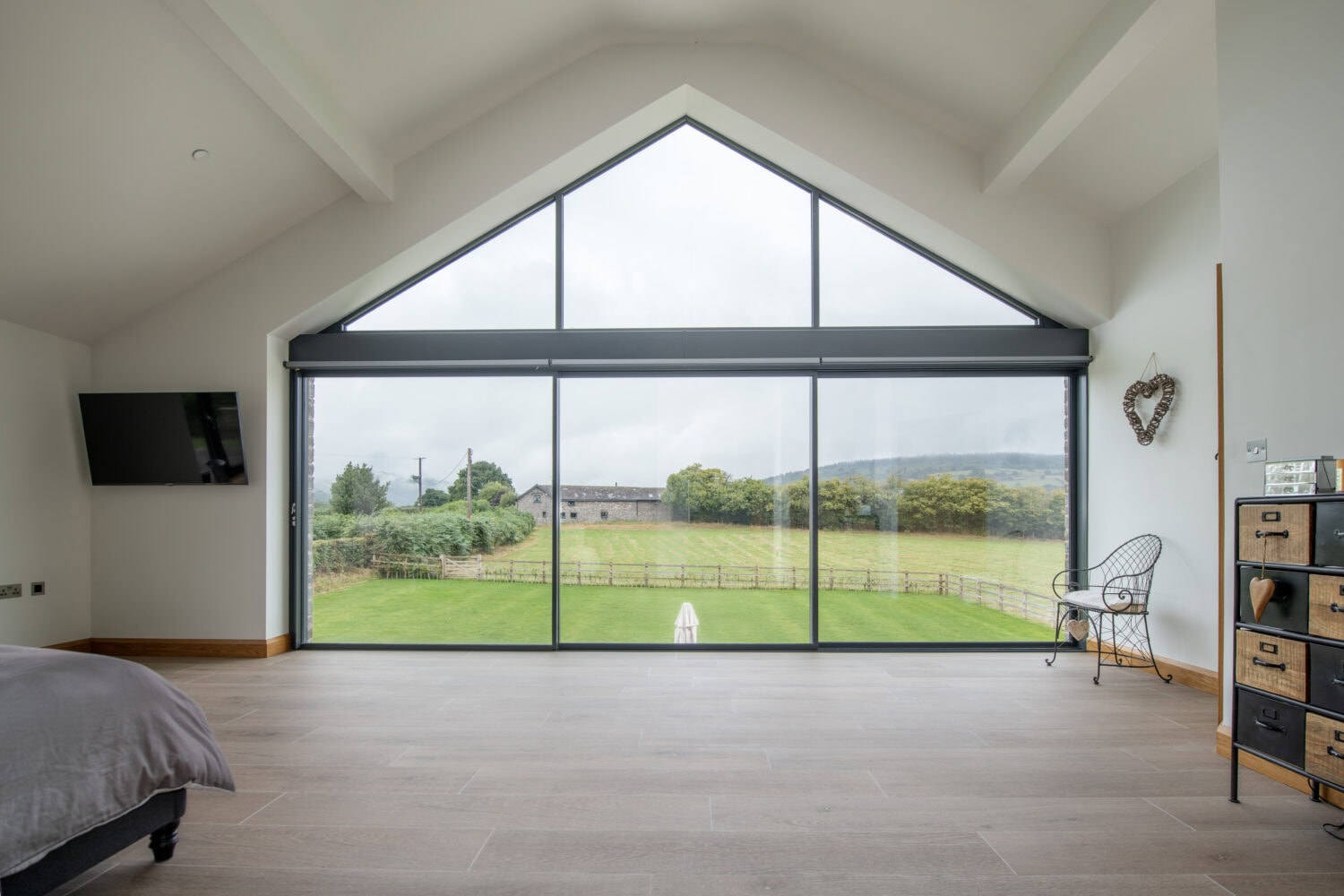 A modern room with bespoke glass solutions featuring large floor-to-ceiling triangular windows overlooking a green field and distant trees, complemented by minimalist furnishings like a bed, chair, dresser, and TV.