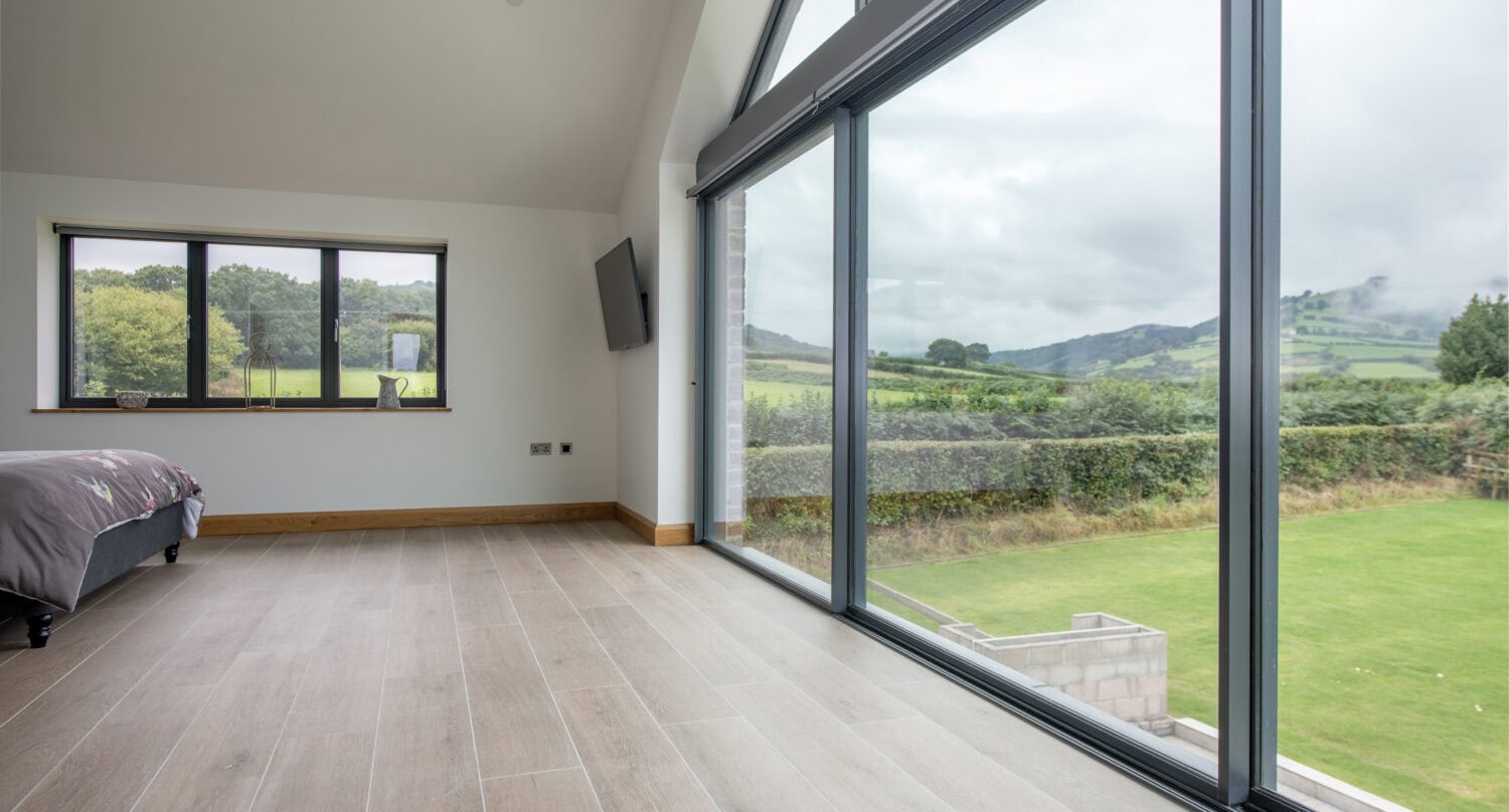 A modern bedroom with large floor-to-ceiling windows and bifold doors opens to a scenic view of green hills and cloudy skies. The room features light wood flooring, a cozy bed in the corner, and minimal furnishings.