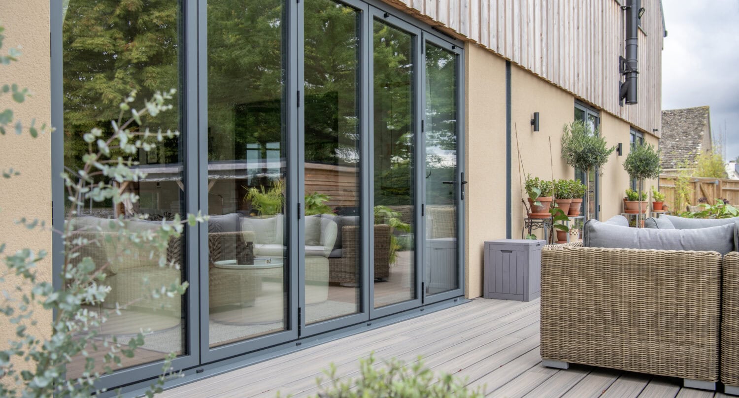 Modern patio with wicker furniture, potted plants, and decking, adjacent to a house with large glass sliding doors and optional glass roofing that reflects lush greenery from the garden.
