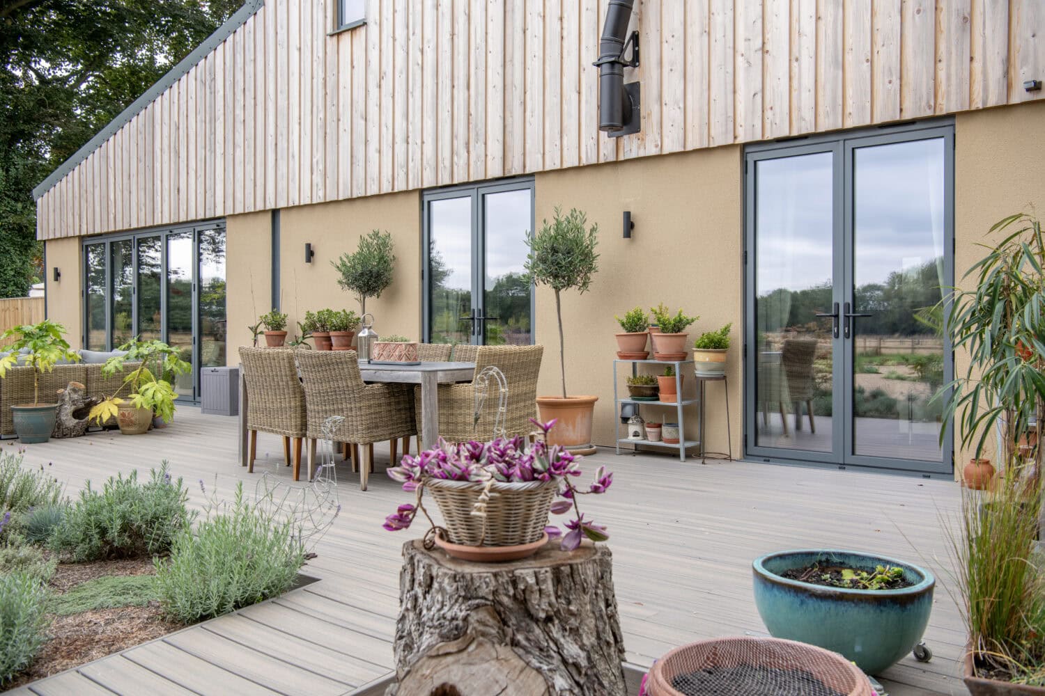 A modern patio with wicker chairs and a wooden dining table, surrounded by potted plants on a wooden deck. Large bespoke glass solutions reflect the garden outside, while a tree stump serves as a unique table.