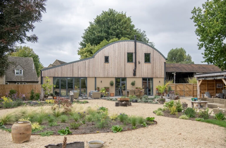A modern barn-style house with a curved wooden roof and large glass windows, featuring bespoke glass solutions and bifold doors, surrounded by a landscaped gravel garden with plants, outdoor seating, and trees in the background.