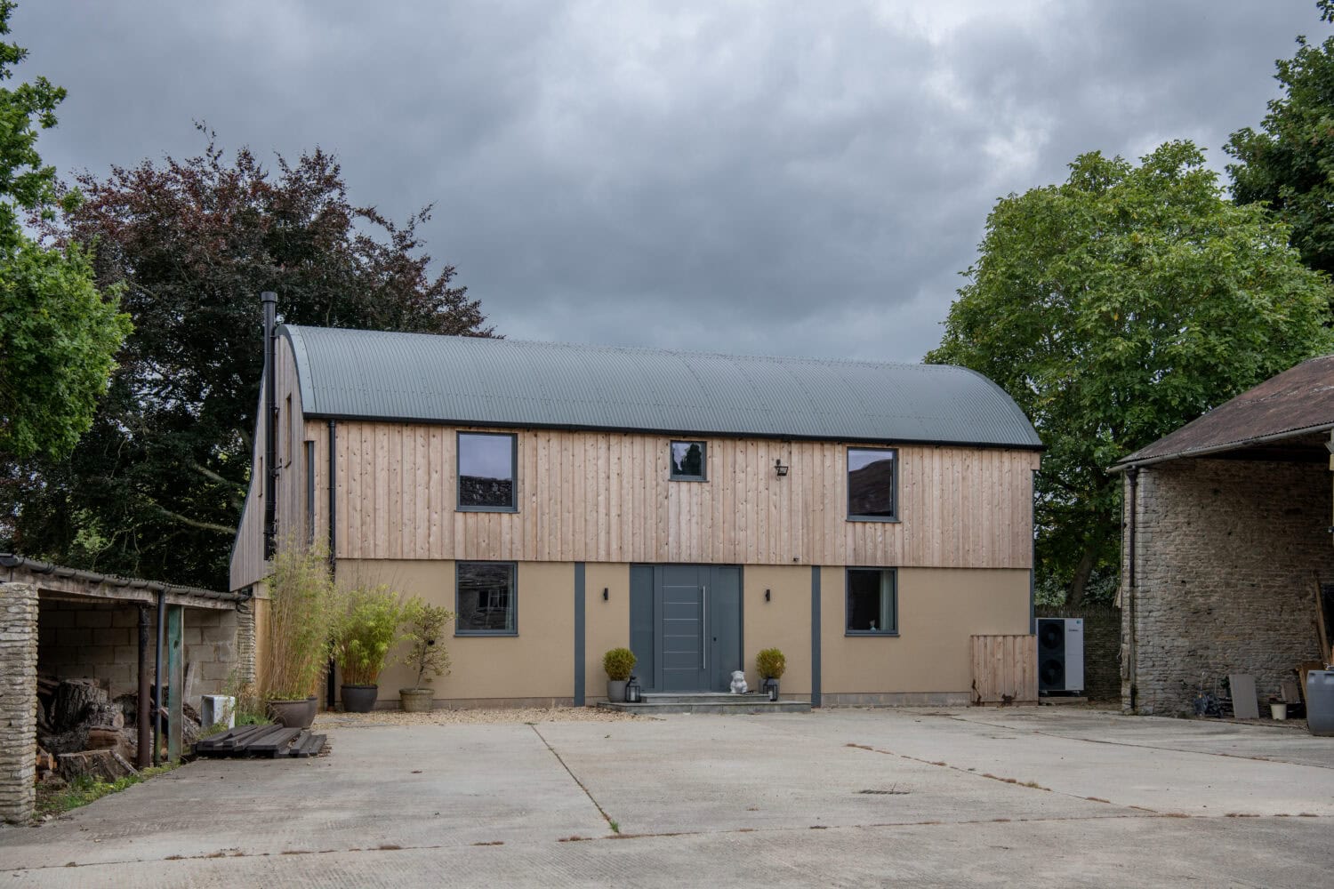 A modern barn-style house with a curved metal roof, wood and beige plaster exterior, two stories, sliding doors, and a double front door. Surrounded by trees, it sits on a concrete courtyard under a cloudy sky.