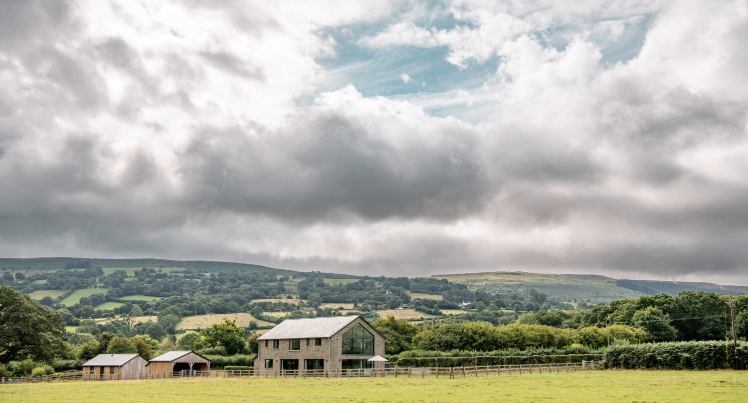 A modern farmhouse with glass roofing and outbuildings sits in a green field, surrounded by fencing and rolling hills under a cloudy sky with patches of blue. Lush trees and hills are visible in the background.