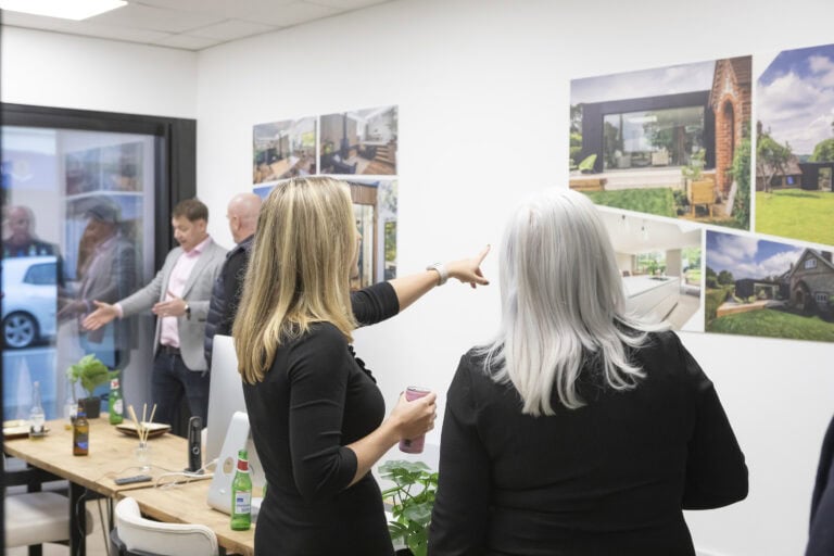 Two women stand in an office, one pointing at interior design photos on the wall near sliding doors. In the background, two men talk by a window. Bottles and plants are on the table in the foreground.