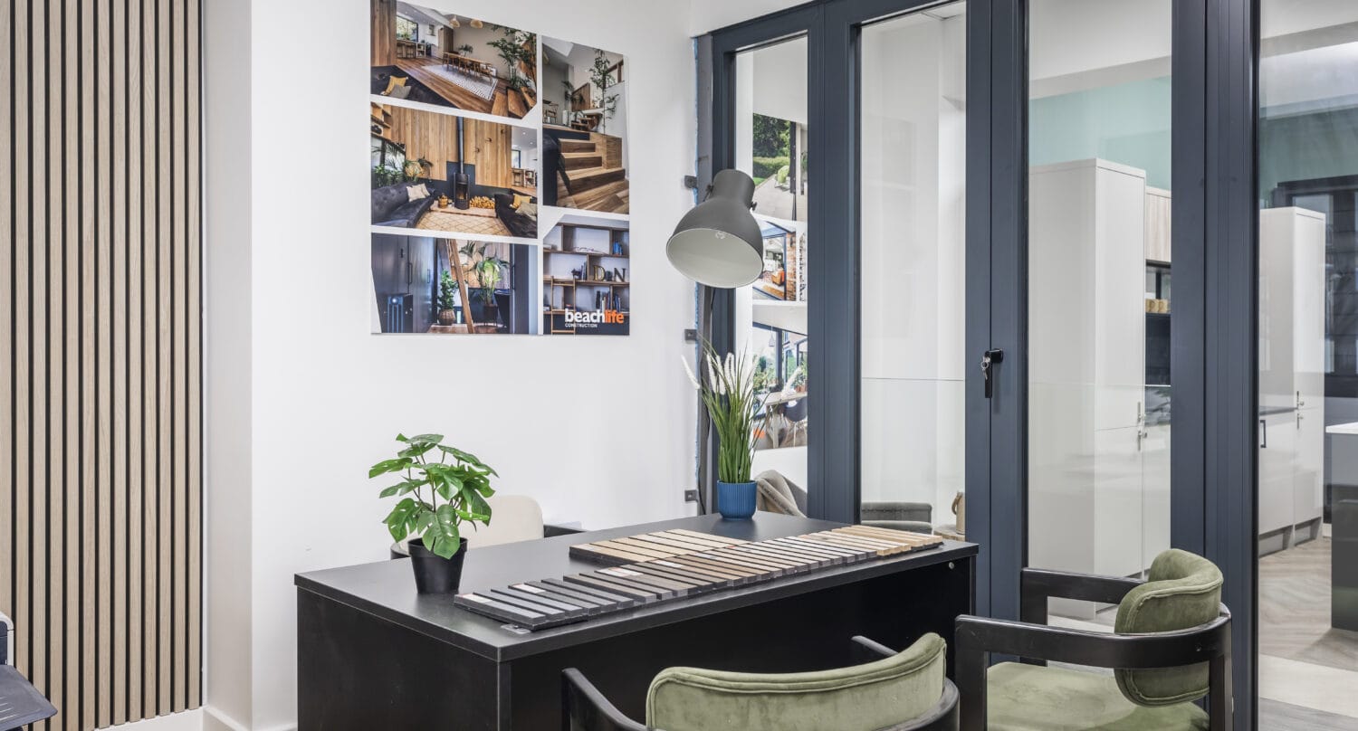 Modern office space with a black desk, two green chairs, a desk lamp, potted plants, and a collage of interior photos on the wall; sleek sliding doors separate the office from another room.