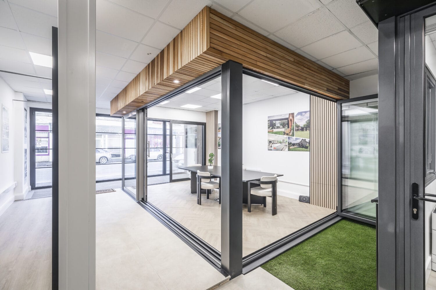 Modern glass-walled office meeting room with bespoke glass solutions, a wooden ceiling accent, four chairs around a table, bright lighting, and large windows with views of parked cars outside.