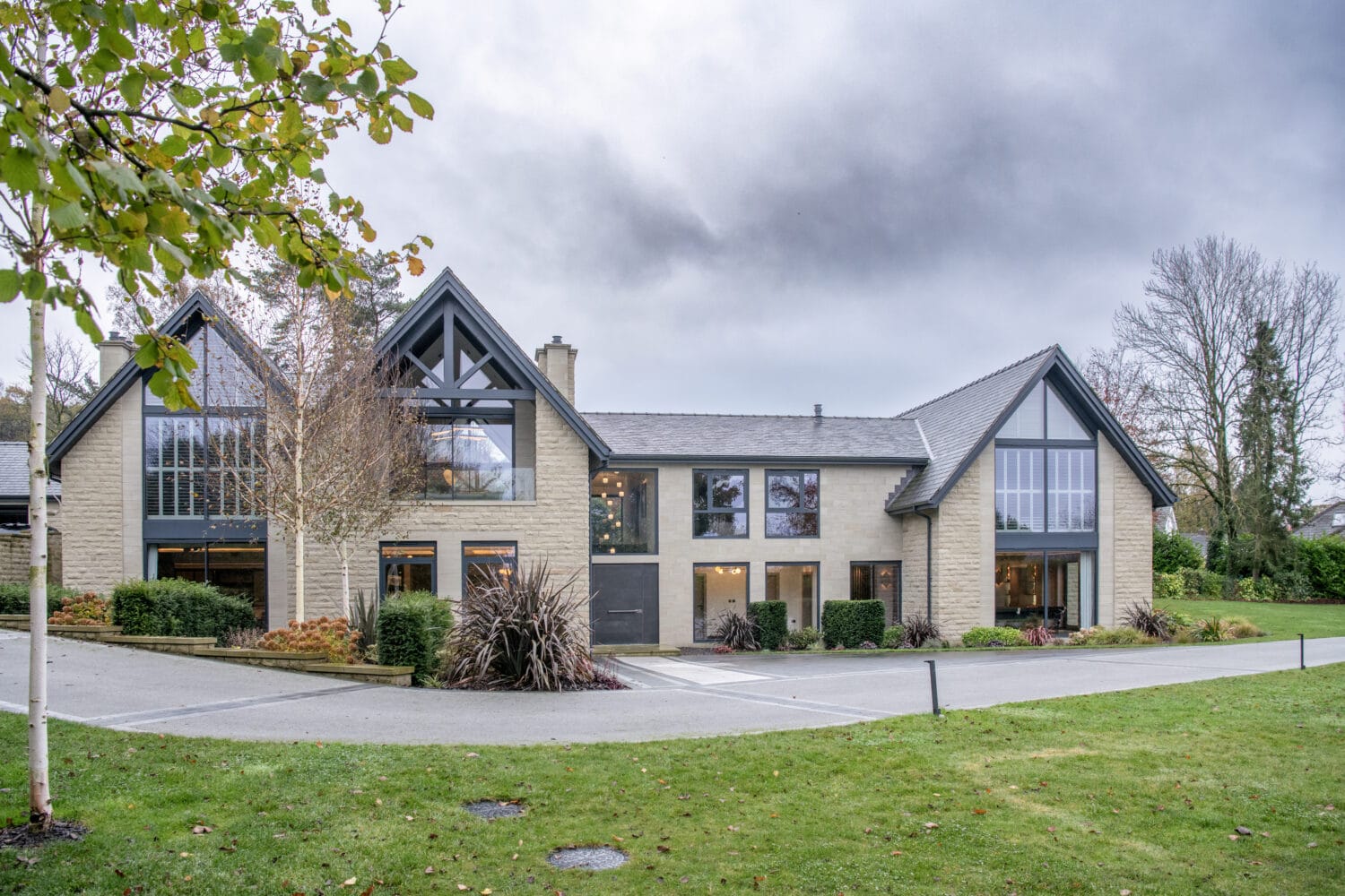 Modern two-story house with large windows, light stone exterior, and gray roof, featuring stylish bifold doors and surrounded by landscaped gardens and trees under a cloudy sky.