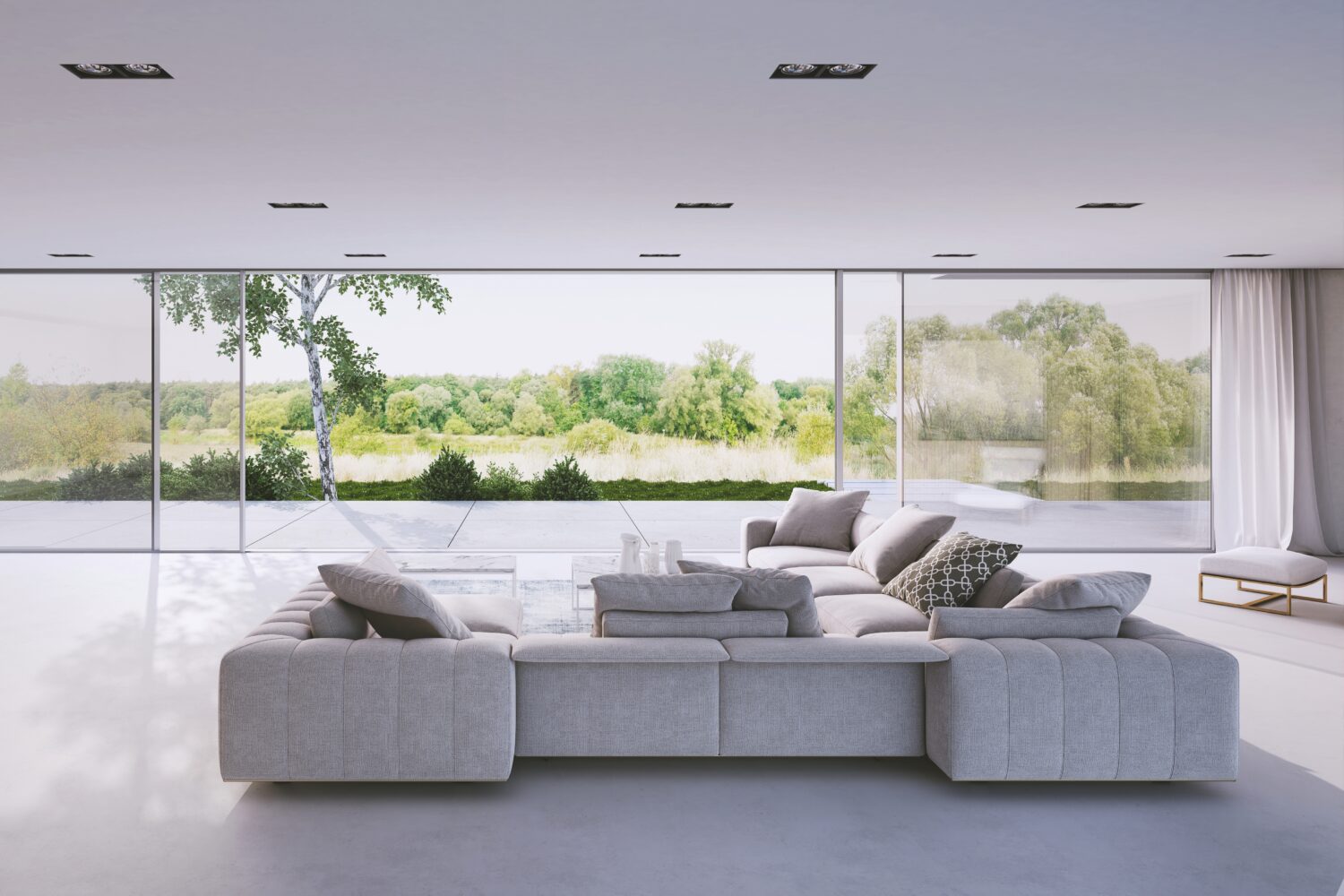 Modern living room with a large gray sectional facing bespoke glass solutions—floor-to-ceiling windows that offer scenic views of green trees. The bright, minimalistic space is filled with natural light and a seamless connection to nature outside.