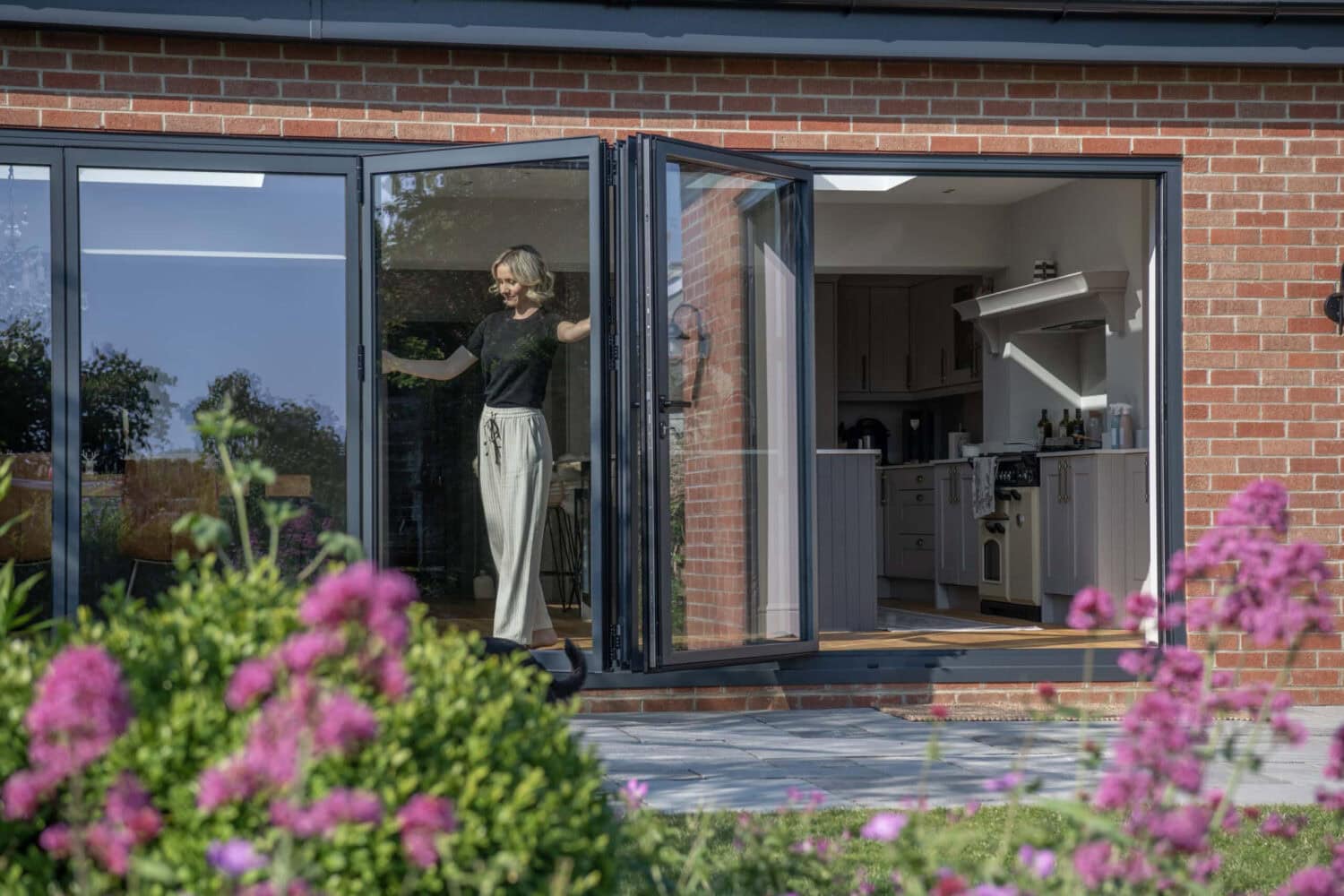 A person stands in an open doorway of a modern brick house with bifold doors, looking outside. Pink flowers and green bushes are in the foreground, and a kitchen is visible inside.