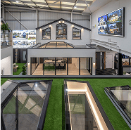A modern showroom displays house designs with large glass windows, skylights, and green artificial grass flooring. Wall-mounted screens highlight bespoke glass solutions for front doors, and the well-lit space features contemporary decor.