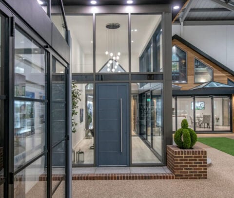 Modern showroom entrance with large glass panels and a sleek gray front door, featuring brick accents and geometric lighting. Decorative plants and contemporary architectural features are visible inside and outside.