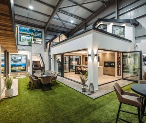 Modern, open-concept showroom with artificial grass flooring, bifold doors, glass walls, dining and seating areas, kitchen display, potted plants, and illuminated ceiling. Stairs and digital screens are visible in the background.