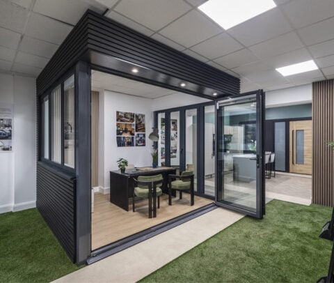 A modern office with glass bifold doors, two green chairs at a desk, and artificial grass flooring leading into the space; bright lighting and contemporary decor create an open, welcoming atmosphere.