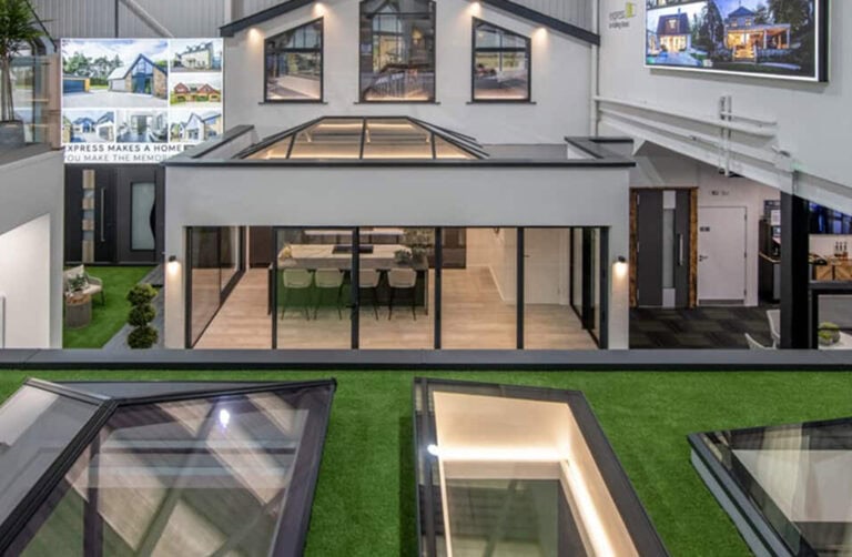 A modern indoor showroom displays model houses with large glass windows, skylights, and bespoke glass solutions. Bright lighting highlights the architecture, while house images are mounted on surrounding walls above the artificial grass.