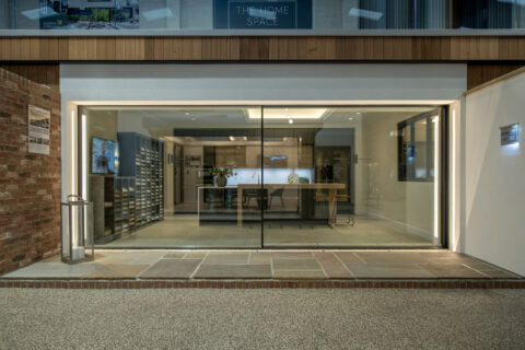 A modern kitchen showroom with large glass windows, bespoke glass solutions, sleek cabinetry, a central island, minimalist decor, and bright interior lighting, viewed from outside on a paved pathway.