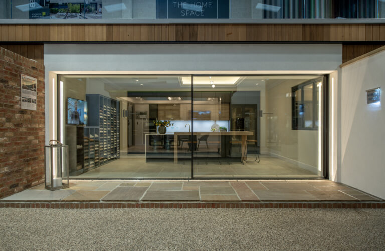A modern kitchen showroom with large glass windows, bespoke glass solutions, sleek cabinetry, a central island, minimalist decor, and bright interior lighting, viewed from outside on a paved pathway.