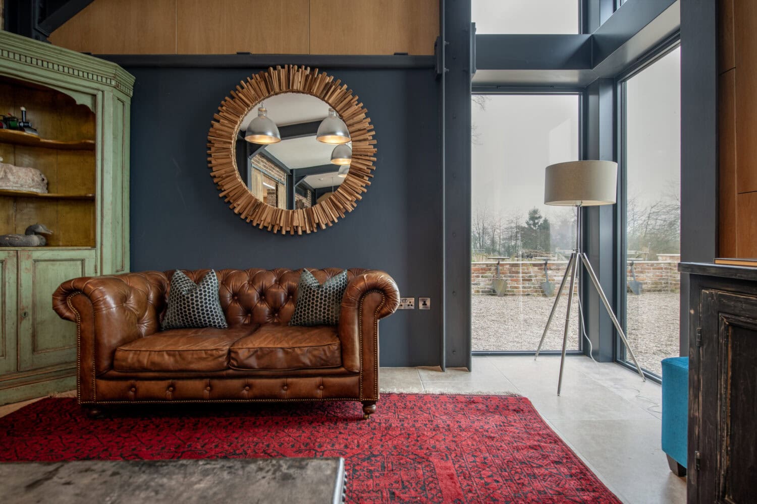 A cozy living room with a brown leather sofa, two patterned cushions, a red rug, a large round wooden-framed mirror, a green cabinet, a floor lamp, and bifold doors that let in plenty of natural light.
