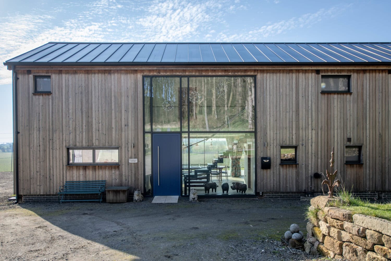 A modern wooden building with a large glass entrance and blue bifold doors, featuring small windows. There’s a bench and plant pots outside, and tree reflections in the glass. The sky is clear with scattered clouds.