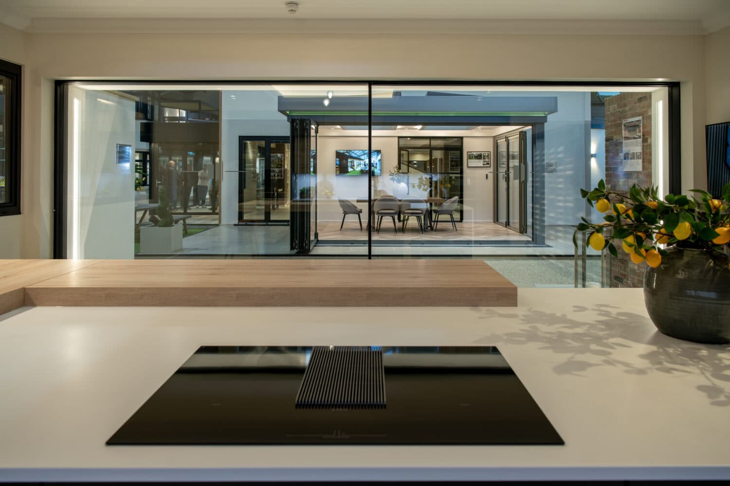 A modern kitchen with a sleek black cooktop on a white island overlooks a spacious dining area and patio through bespoke glass sliding doors. A vase with green and yellow flowers sits on the counter, enhancing the elegant ambiance.