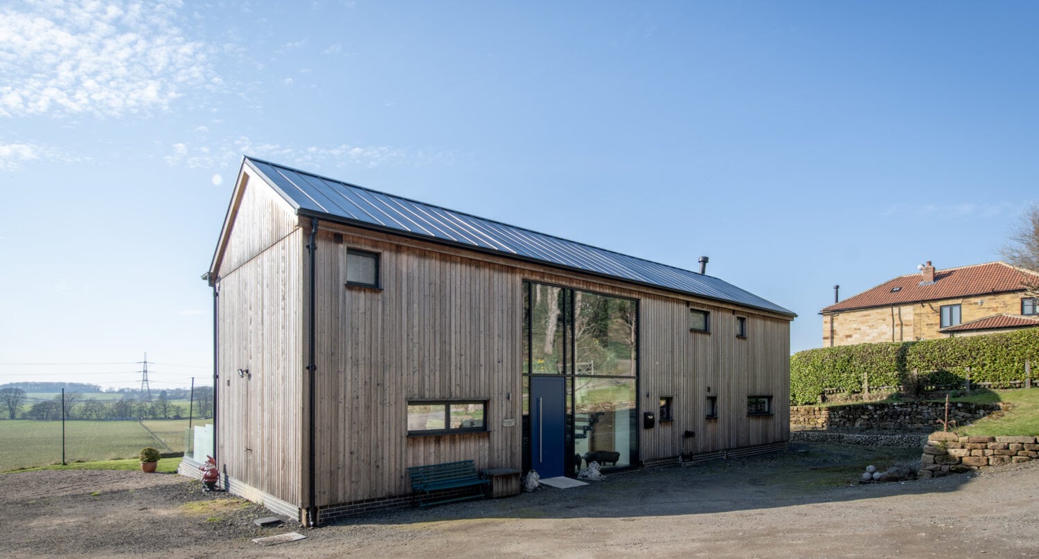 A modern, rectangular wooden house with a metal roof and large central bespoke glass solutions, including sliding doors, stands on a dirt yard surrounded by fields and a stone house in the background under a clear blue sky.