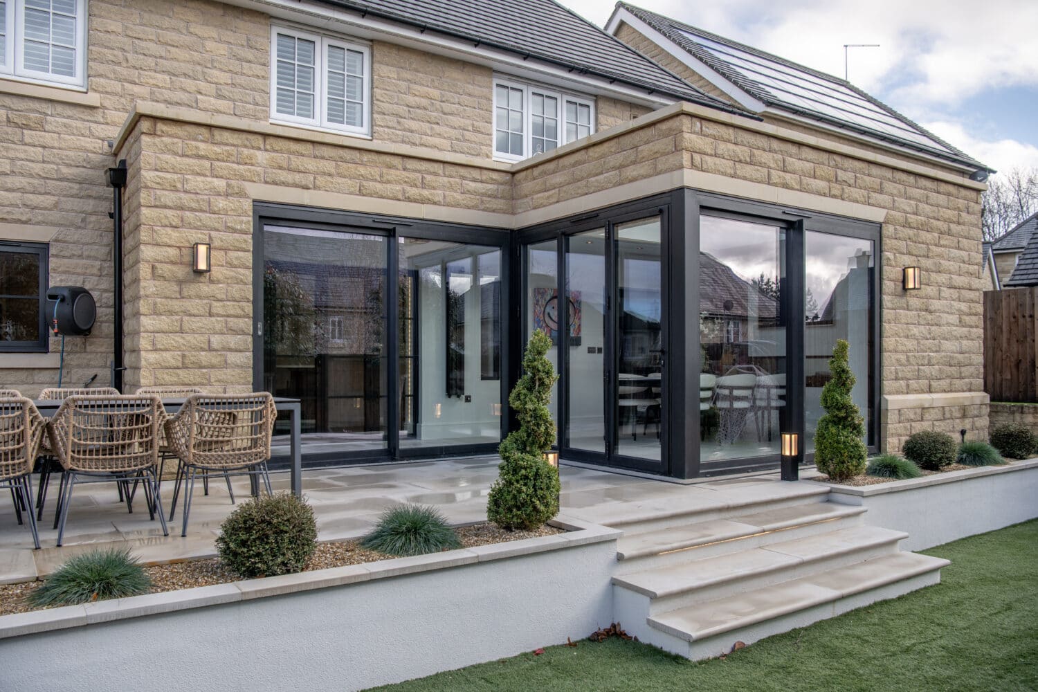 Modern house with light stone exterior, bespoke glass solutions including large black-framed sliding doors, a patio with outdoor dining set, potted shrubs, manicured lawn, and steps leading up to the entrance.