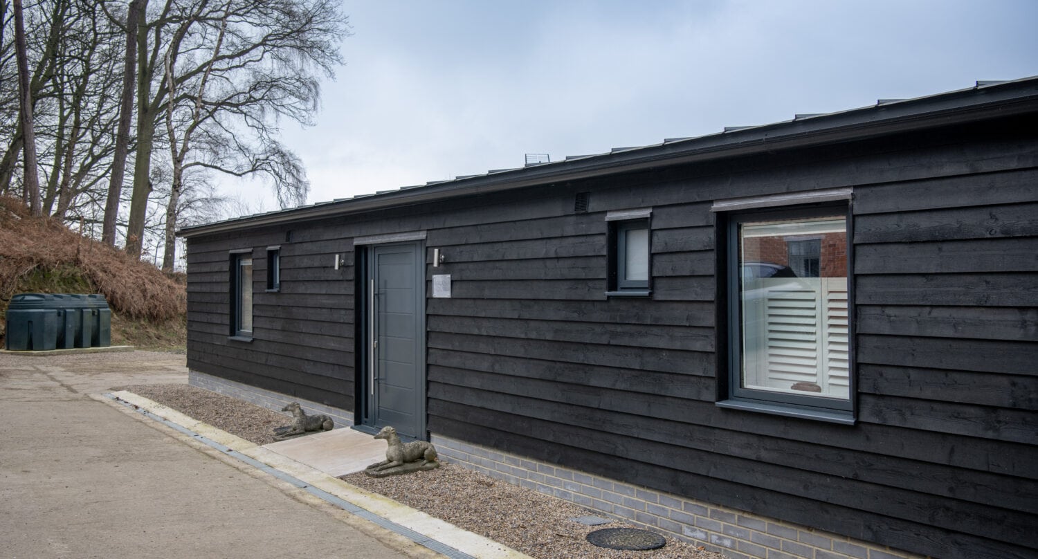 Modern single-story black wooden building with large windows, a gray front door, brick foundation, two small dog statues by the entrance, and leafless trees in the background on a cloudy day—featuring bespoke glass solutions throughout.