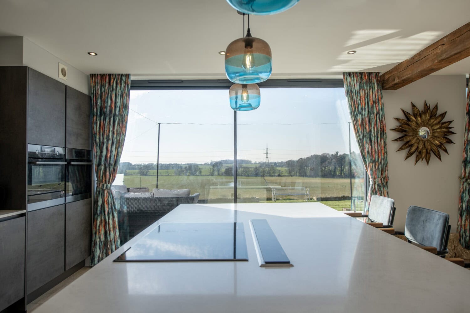 Modern kitchen with a white island countertop, built-in ovens, colorful patterned curtains, and large bifold doors overlooking a scenic outdoor landscape. Blue hanging lights and a sunburst wall decor brighten the space.