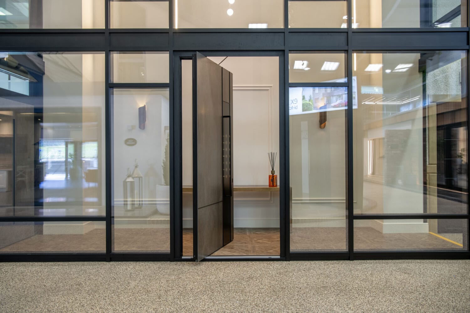 Glass wall with a large modern pivot front door, partially open, revealing a minimal interior with a wooden console table and orange decorative sticks. The speckled floor and bright lighting enhance these bespoke glass solutions.