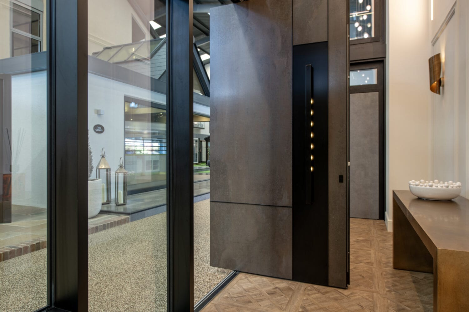 A modern entryway with a large pivoting front door, glass walls, a bench with a decorative bowl, and a view of an outdoor courtyard with lanterns and greenery beneath sleek glass roofing.
