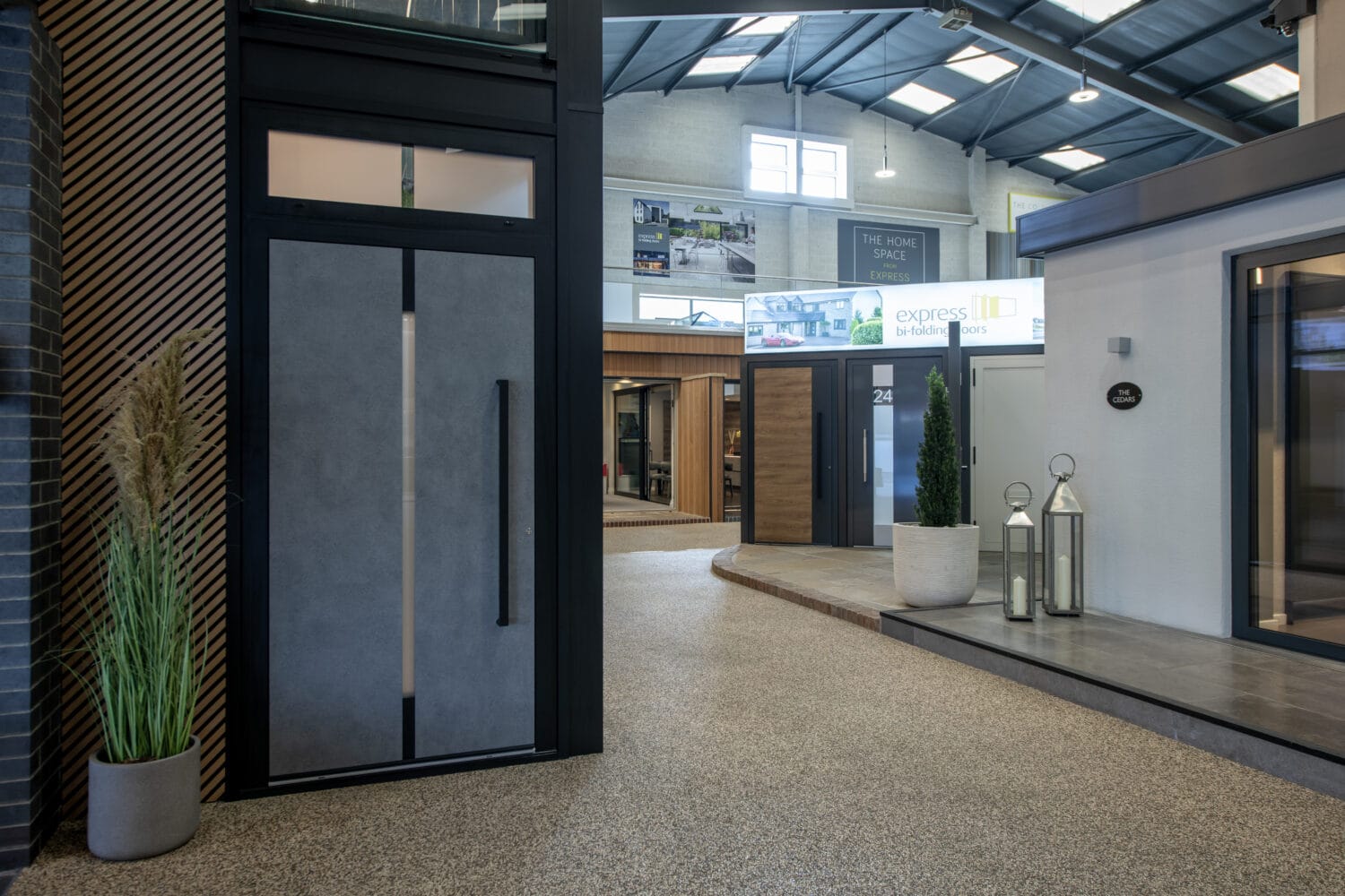 A modern indoor showroom displays various contemporary front doors and windows, with plants and decorative lanterns near the entrances under a high, industrial-style ceiling featuring sleek glass roofing.