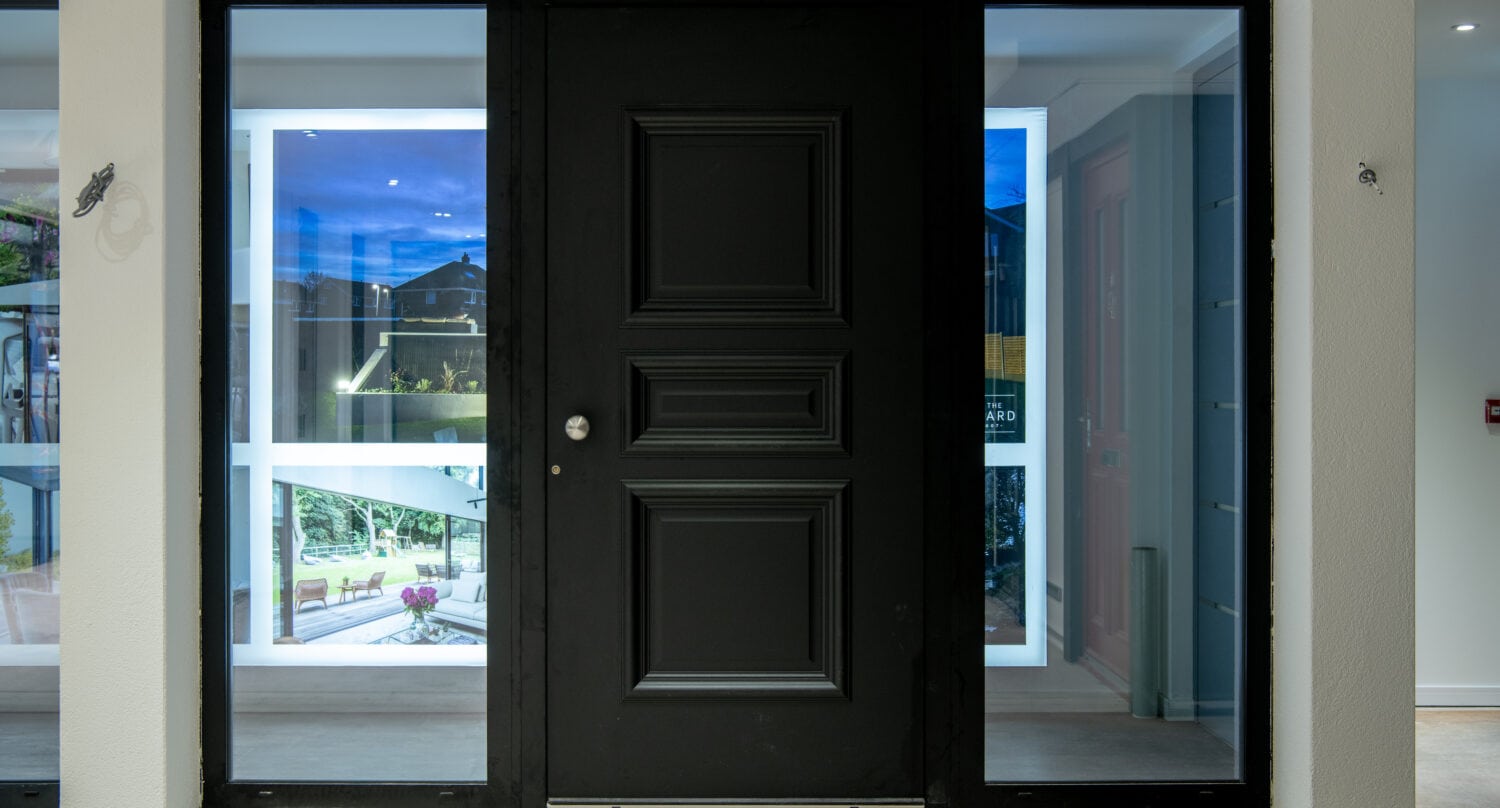 A modern black front door with decorative panels, flanked by tall glass side panels, opens into a contemporary home interior with white walls and large windows, showcasing bespoke glass solutions and outdoor greenery.