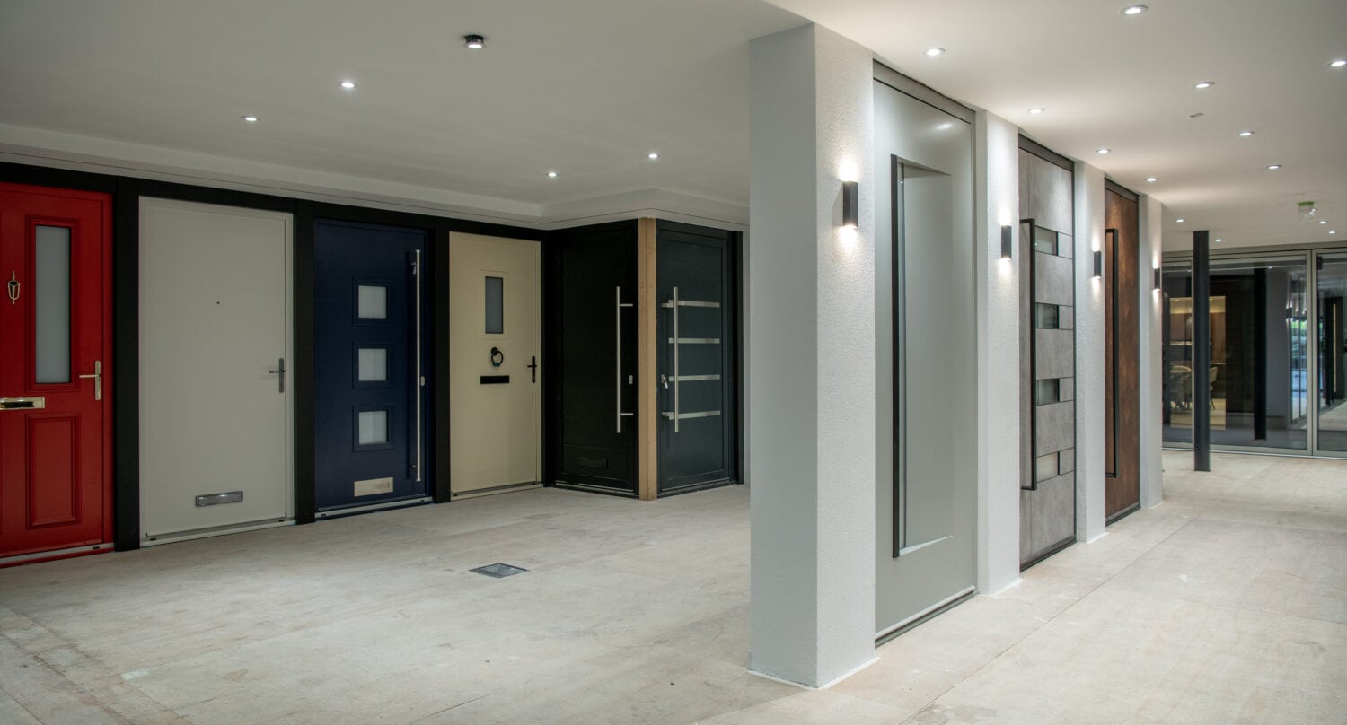 A showroom displaying various front door styles in different colors and designs, mounted side by side on a spacious floor with modern lighting and sleek sliding doors, all under a bright, glass roofing that fills the area with natural light.