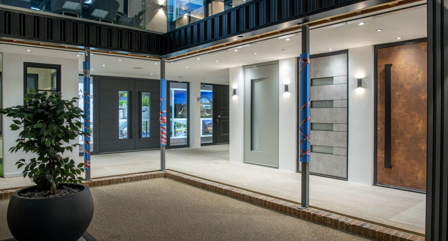 Modern building entryway with large glass windows showcasing bespoke glass solutions, minimalist lighting, gray and brown wall panels, and a potted plant in the foreground. Reflections reveal a bright and open interior space.